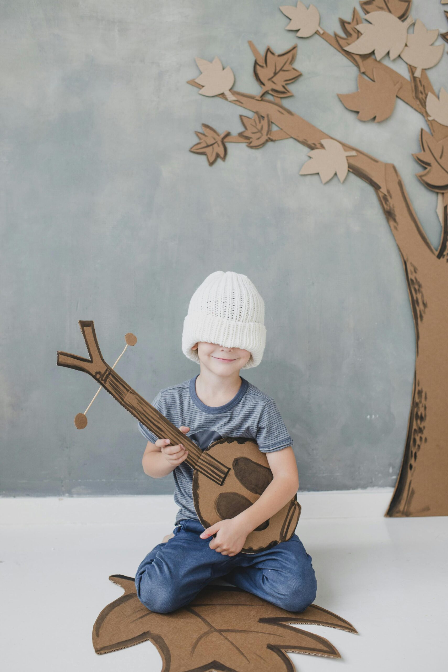 Free Full body smiling cute little boy in casual clothes with eyes covered with white knitted hat sitting on floor with toy dombra against gray wall and carton fake tree Stock Photo