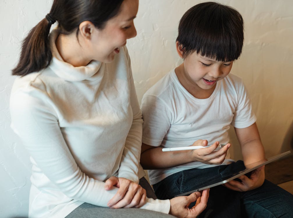 Free From above of crop glad mother in casual clothes sitting near cheerful ethnic boy with pen and tablet while using social media in house Stock Photo