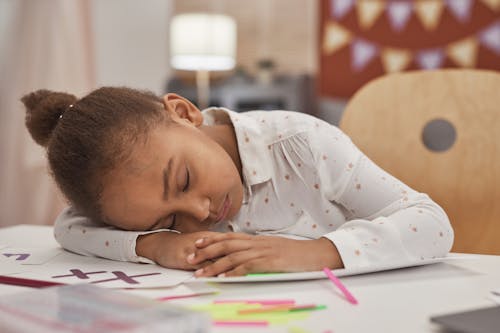 Free A tired young girl with closed eyes rests her head on homework at a table indoors. Stock Photo