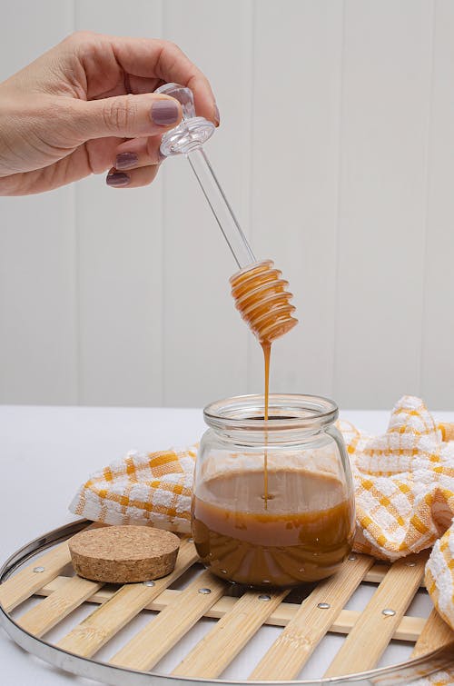 Free Close-up of a hand with manicured nails holding a honey dipper over a glass jar with cork lid, placed on a checkered cloth. Stock Photo