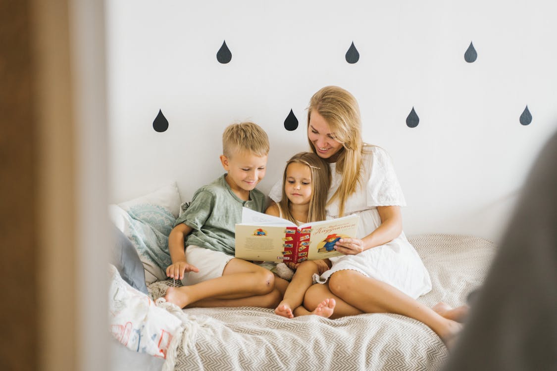 Free A joyful mother reads a storybook to her young children while sitting on a bed. Stock Photo