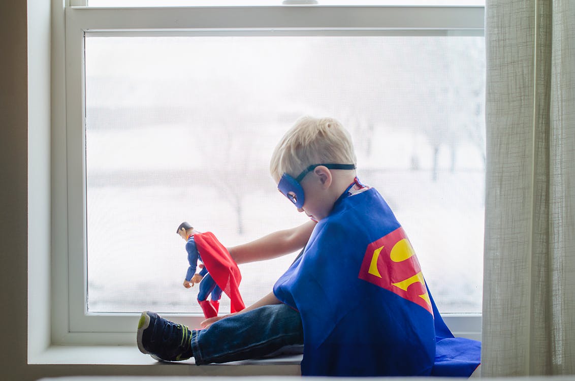 Free A young boy dressed as a superhero, playing with a toy by a window, embracing his imagination. Stock Photo