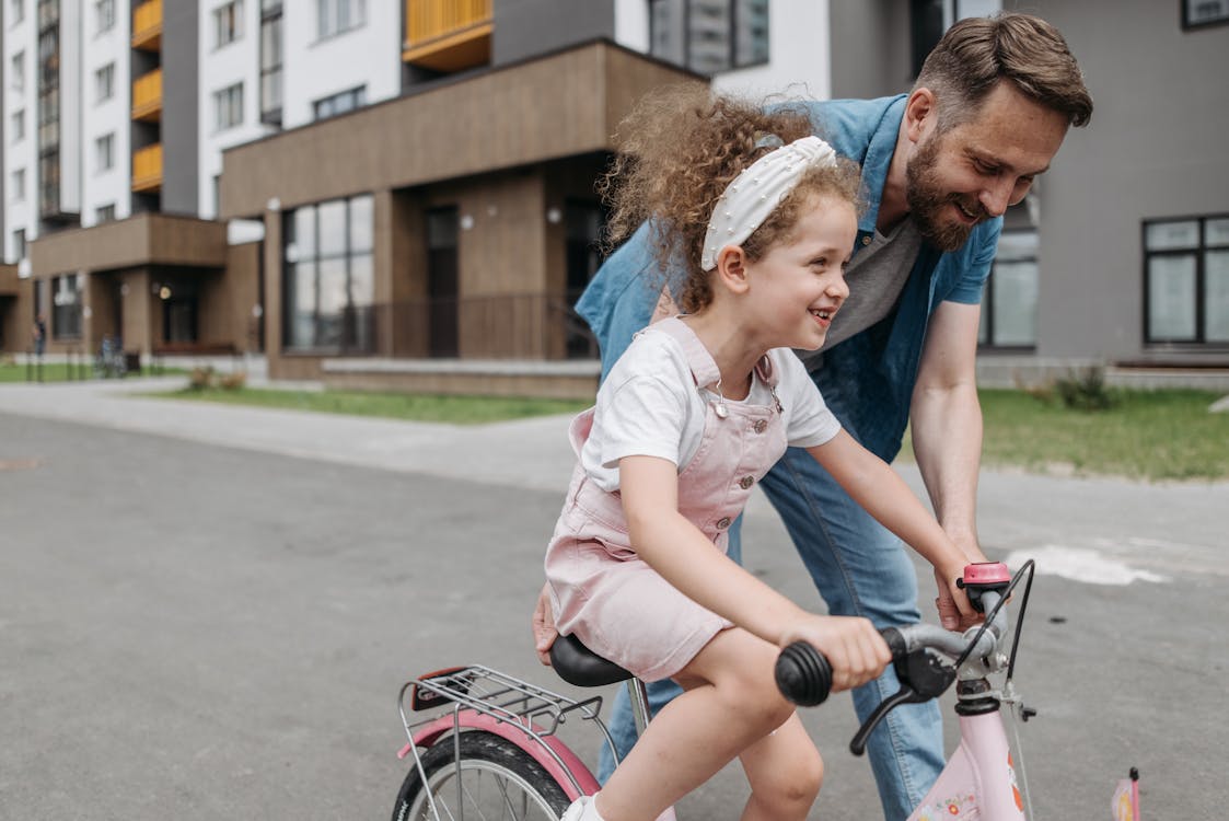 Free Father helps daughter learn to ride a bike in an urban setting, capturing joy and bonding. Stock Photo