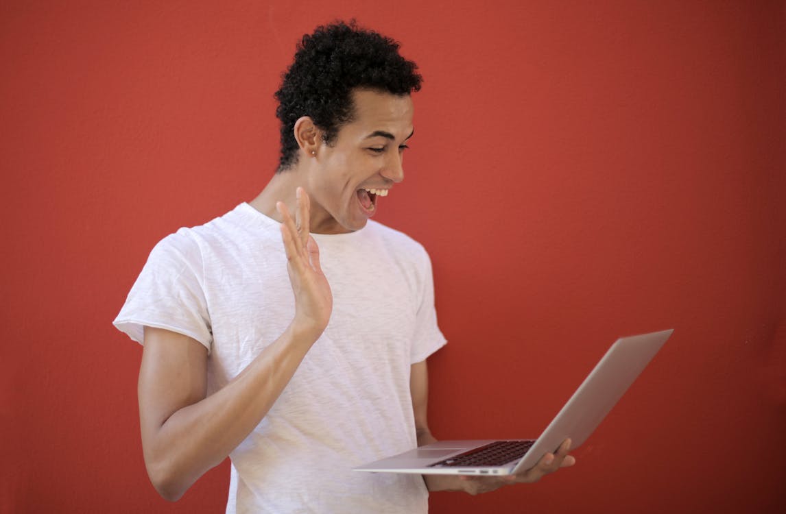 Free Young man making a cheerful video call on a laptop with a red background, expressing joy and surprise. Stock Photo