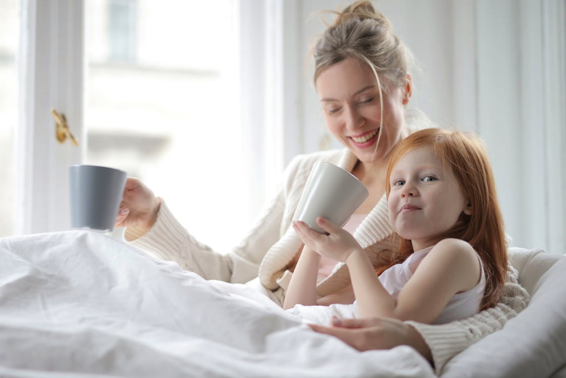 Free A joyful mother and daughter share a cozy morning with warm drinks in a bright bedroom. Stock Photo