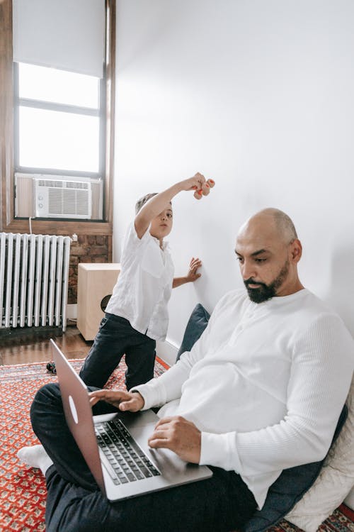 Free A father works on his laptop while spending quality time with his son in a cozy room. Stock Photo